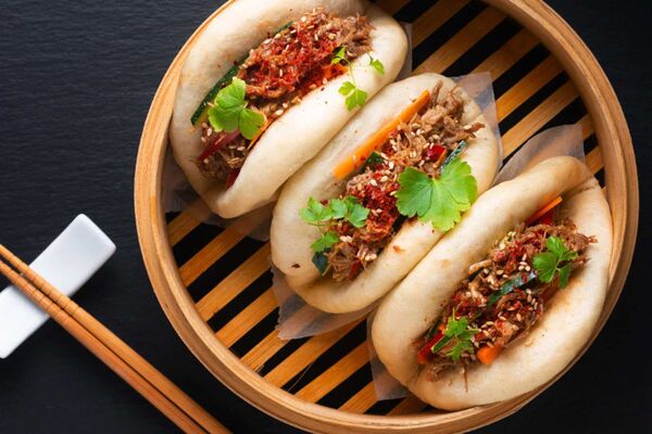 Drei frische Bao Buns in einer Bambus-Dämpf-Schale angerichtet, garniert mit Koriander. Links daneben liegen Ess-Stäbchen.